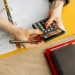 Tangan menggunakan kalkulator di meja dengan dokumen akuntansi dan folder keuangan